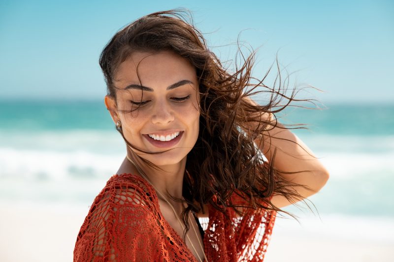 Latin fashion woman wearing red lace dress at beach and enjoy fresh breeze. Portrait of carefree tanned girl relaxing at beach during summer vacation. Young smiling beauty woman during summer vacation.